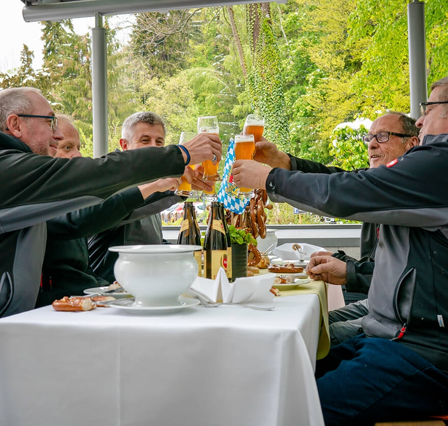 Eine Gruppe aus fünf Herren sitzen um einen mit Weißwürsten, Brezen und Bieren gedeckten Tisch und stoßen mit einem vollen Glas alkoholfreiem Weißbier an.