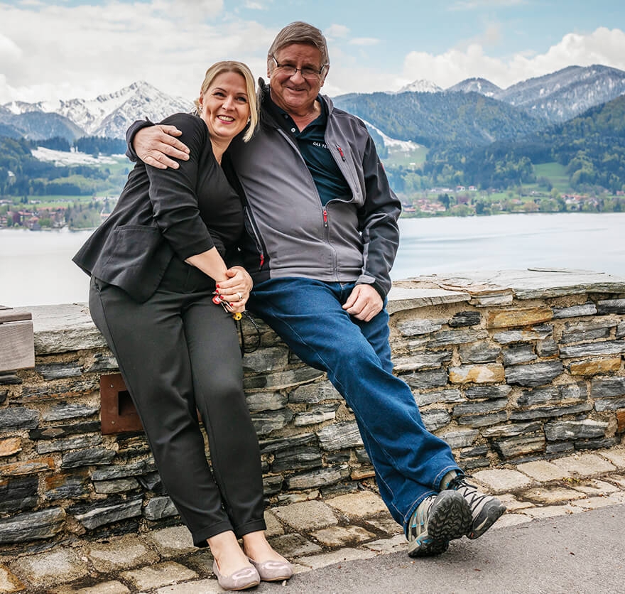 Eine Frau und ein Mann sitzen auf einer niedrigen Mauer direkt am Tegernsee mit Blick auf die Berge im Hintergrund.