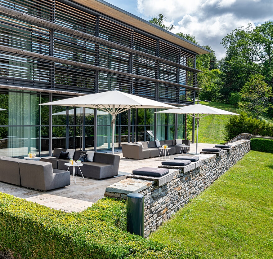 Außenaufnahme vom Hotel Das Tegernsee mit einer langen Terrasse vorm Gebäude, auf der mehrere Lounge-Stühle und Sonnenschirme aufgebaut sind.