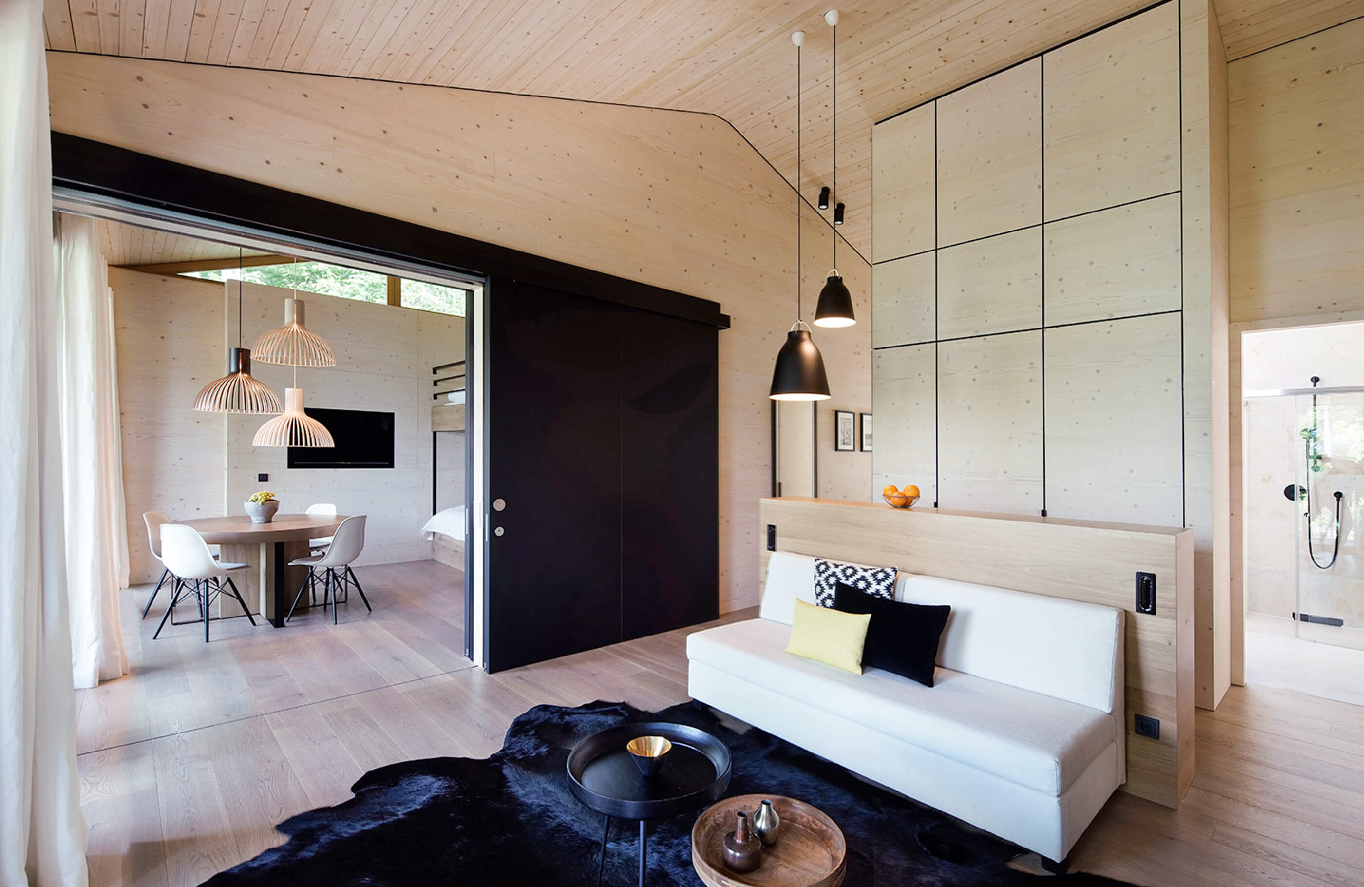 Aufnahme im Wohnraum des Alpenchalets vom Lifestyle Hotel in Bayern am Tegernsee mit einem schwarzen Teppich, weißem Sofa und Blick ins angrenzende Zimmer mit einem Essbereich bestehend aus einem runden Holztisch und vier weißen Stühlen.