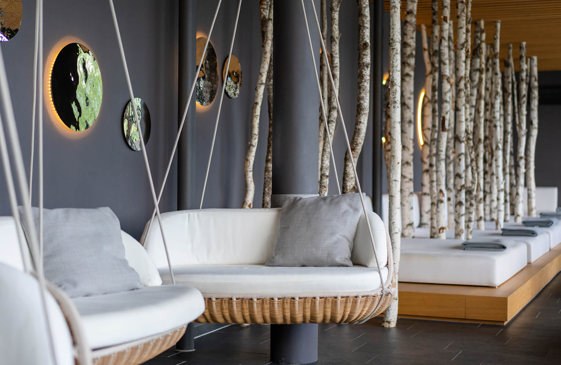 Aufnahme im Ruheraum des Spa-Bereichs im Hotel Das Tegernsee mit hellgrauen Wänden, mehreren Ruheliegen, dekorativen Birkenbaumstämmen dazwischen und im Vordergrund zwei Lounge-Hängesessel.