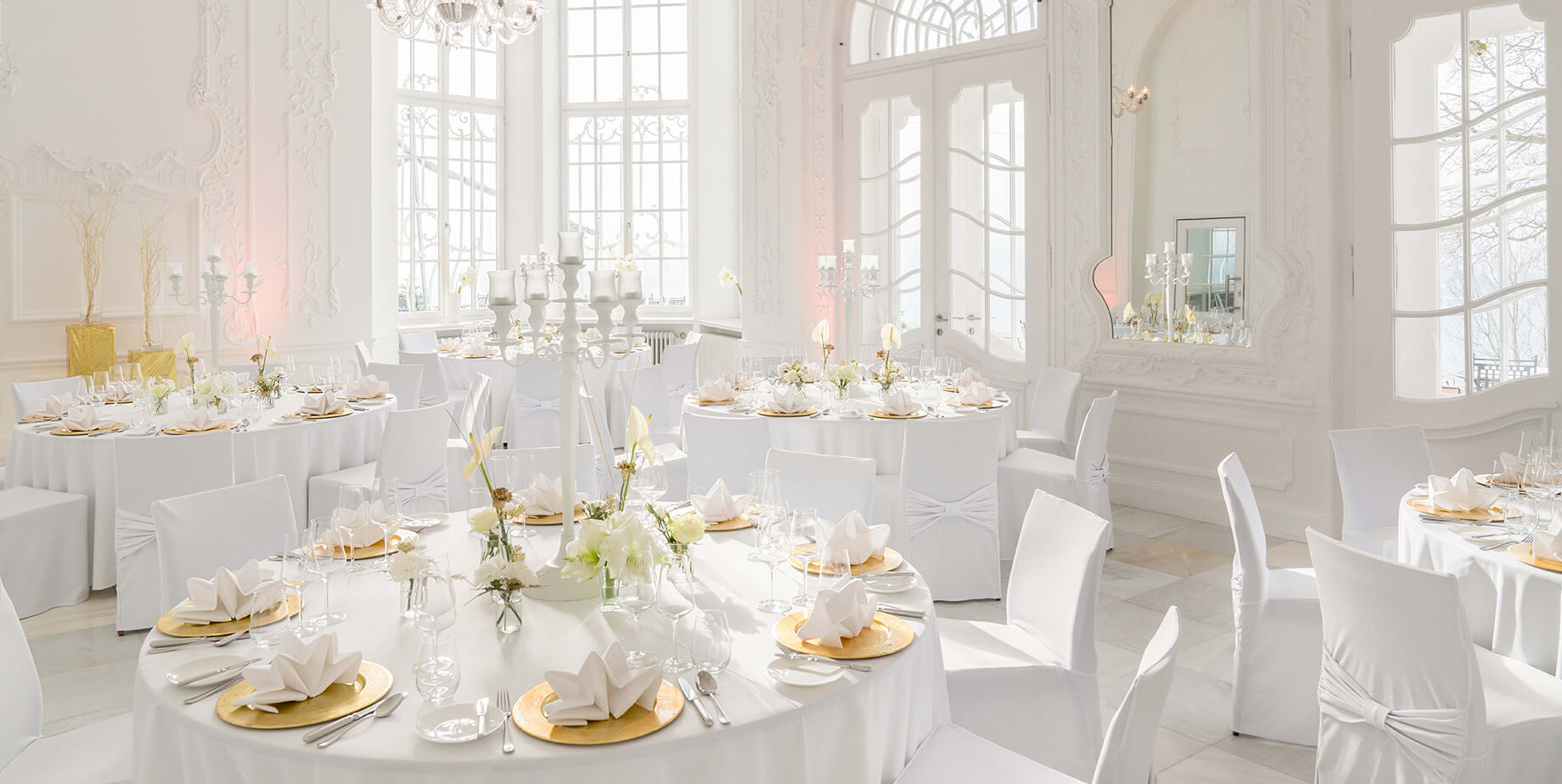 Aufnahme eines Festsaals mit hohen Wänden und Bogenfenstern in der Hochzeitslocation am Tegernsee mit mehreren in weiß und Gold eingedeckten Rundtischen und Stühlen mit weißen Stuhlhussen.