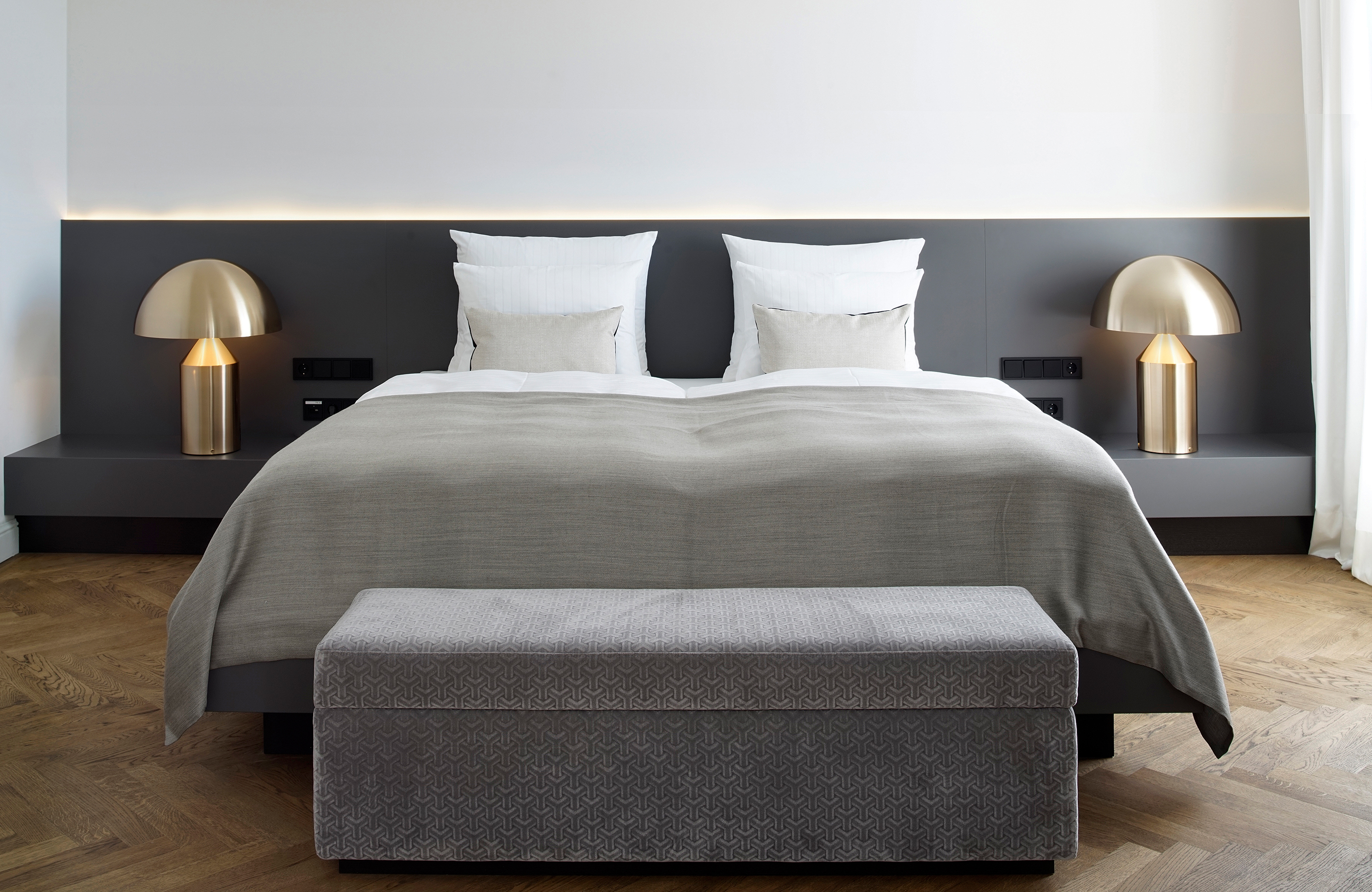 Blick in ein Doppelzimmer im Hotel Das Tegernsee mit einem großen, in weiß-grauen Farben bezogenem Bett, vor dem eine graue Truhensitzbank steht.