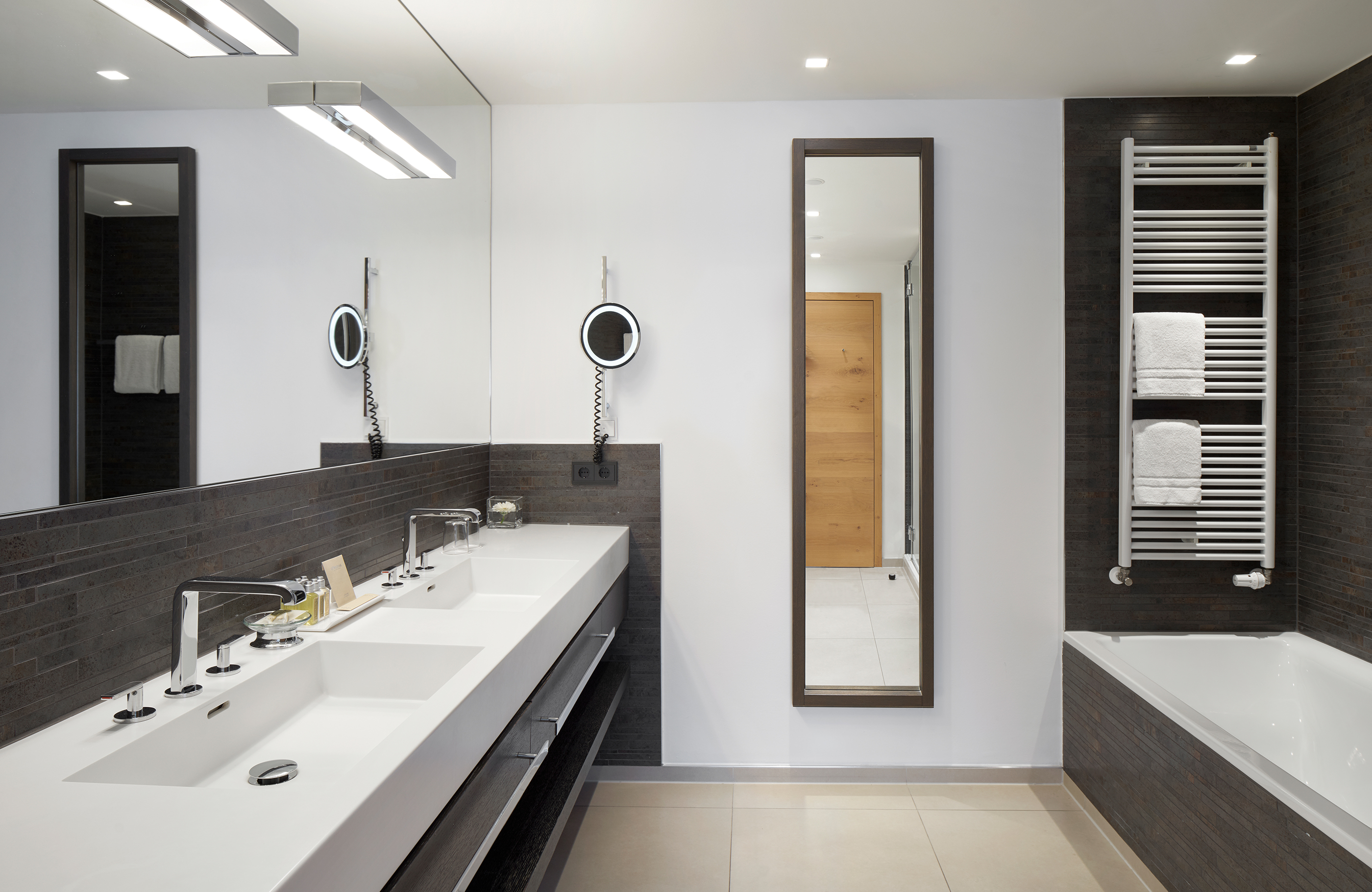 Aufnahme im Badezimmer mit einem doppelten Waschbecken, Badewanne, großen Spiegeln und dunklen modernen Fliesen