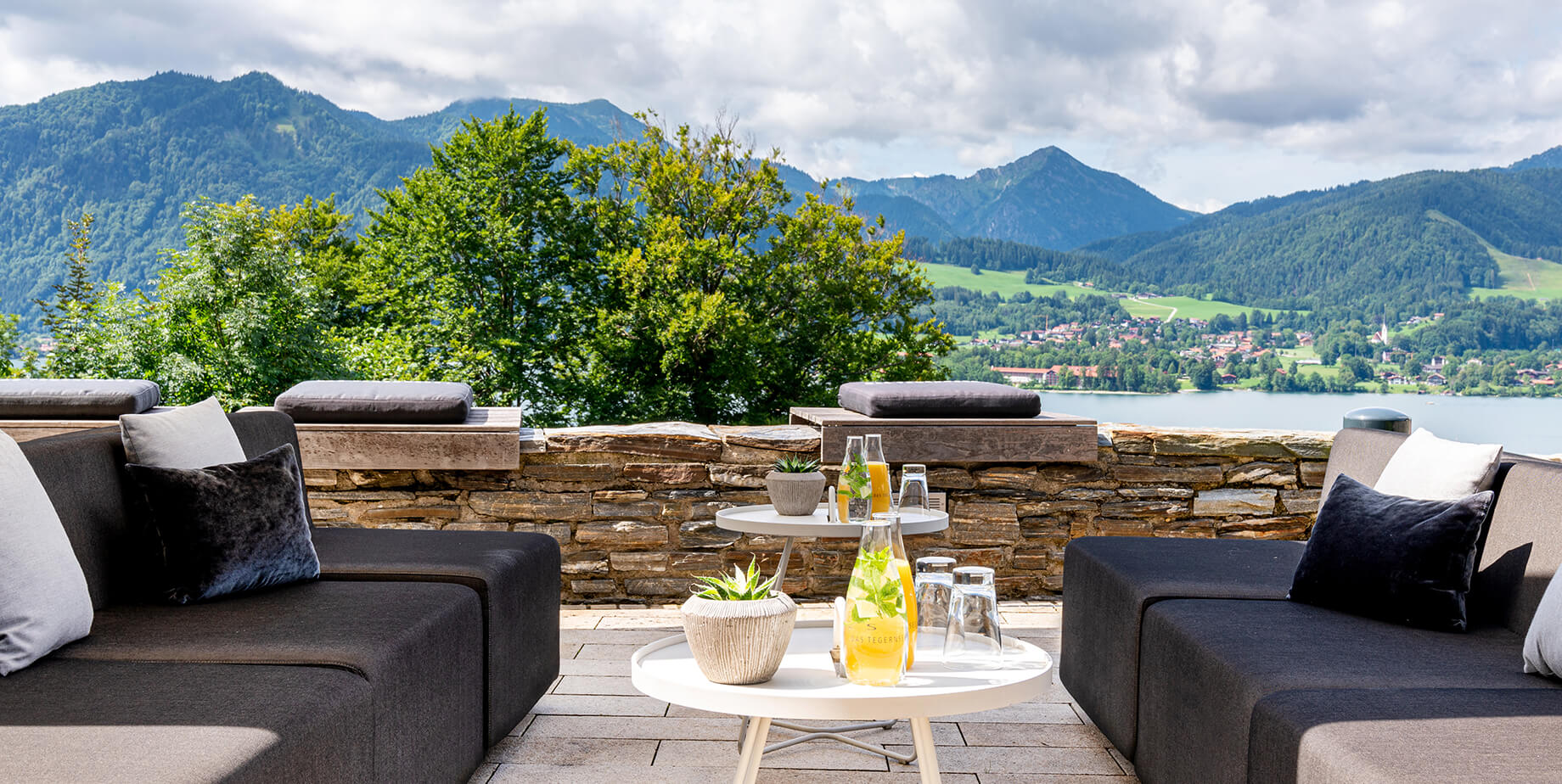 Aufnahme einer Terrasse mit mehreren hell- und dunkelgrauen Lounge-Sesseln im Hotel Das Tegernsee mit Panoramablick auf den See und die Berge.