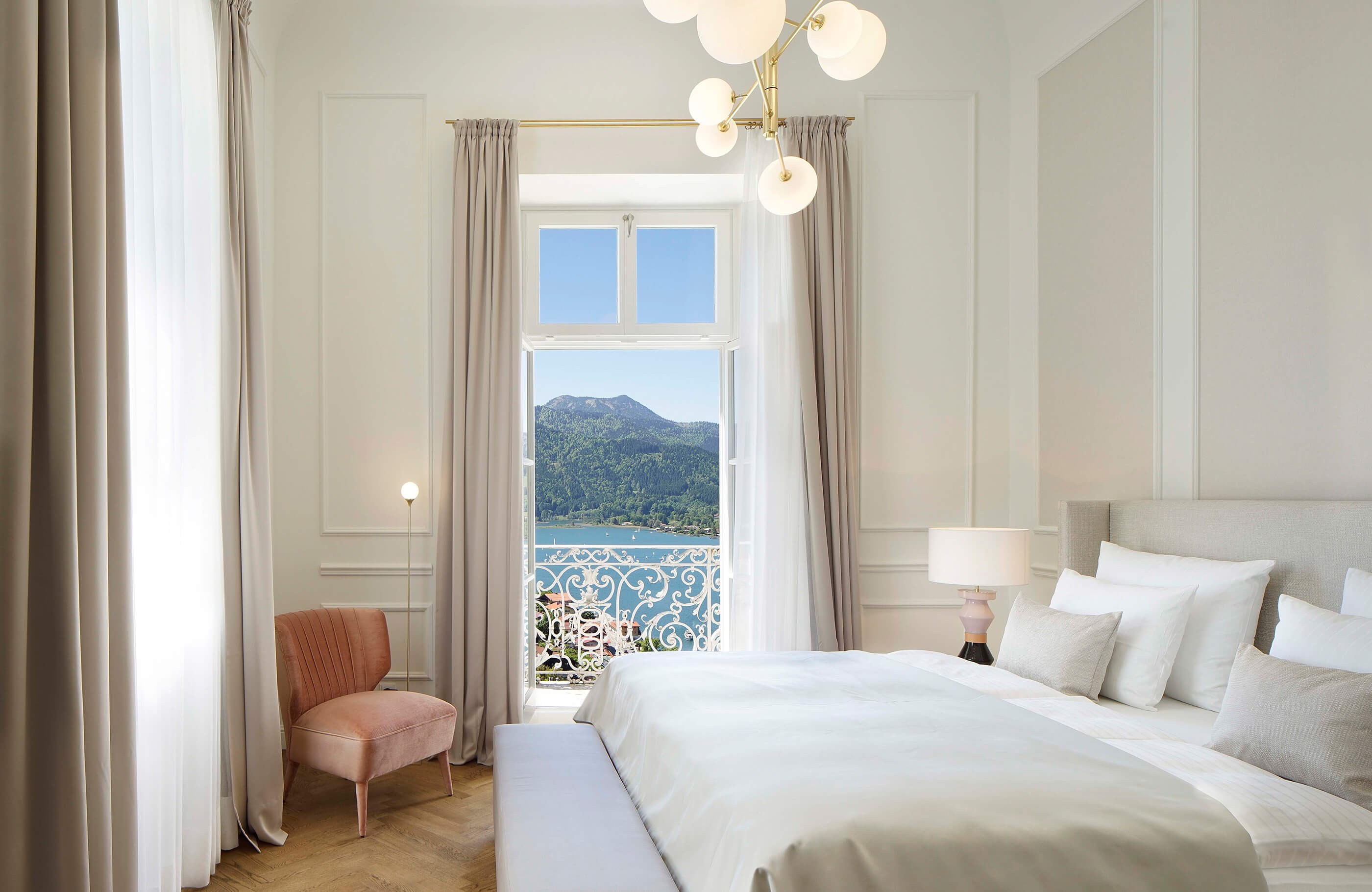 Blick in das Schlafzimmer einer Deluxe Suite im Hotel Das Tegernsee mit einem großen, weiß bezogenem Doppelbett, langen hell-grauen Vorhängen vor zwei Fenstern und Zugang zum Balkon mit Seeblick.