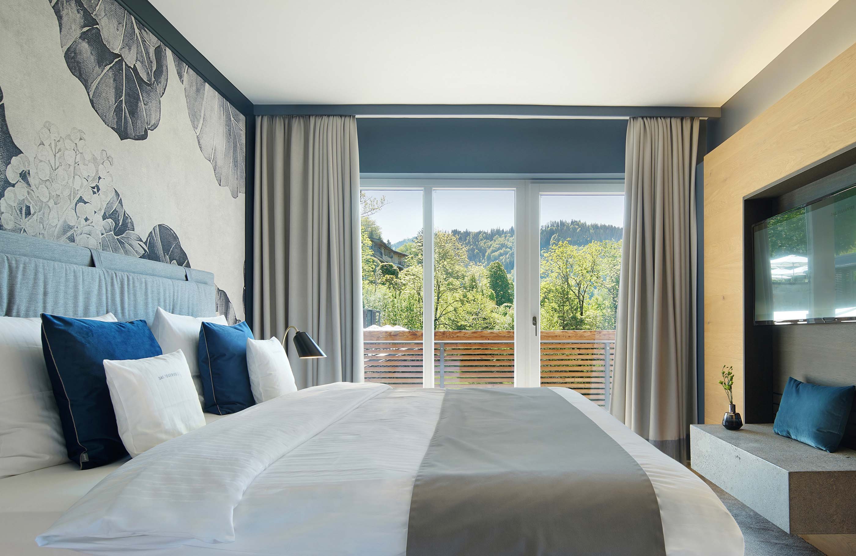 Blick über ein großes Bett mit blauen Akzenten und einer großen Fensterfront