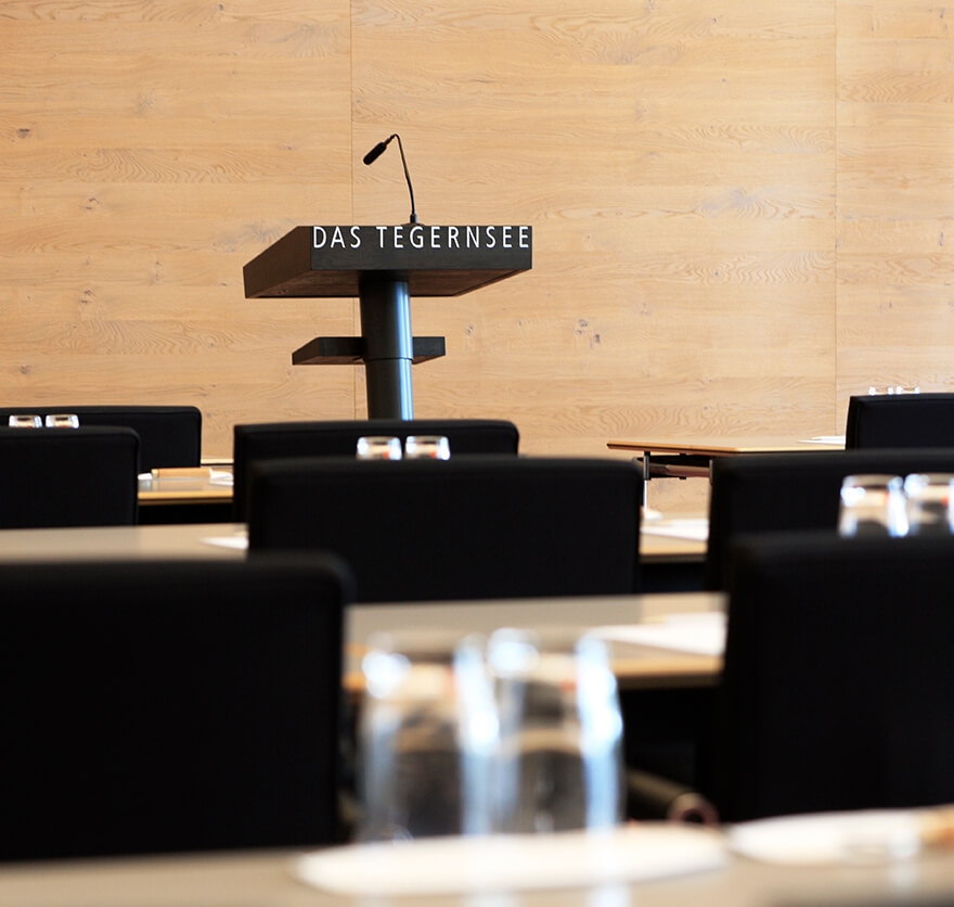 Aufnahme in einem Tagungsraum mit Tischen, die in Reihen mit schwarzen Stühlen angeordnet sind und im Zentrum im Bildhintergrund befindet sich ein Rednerpodest mit Mikrofon.