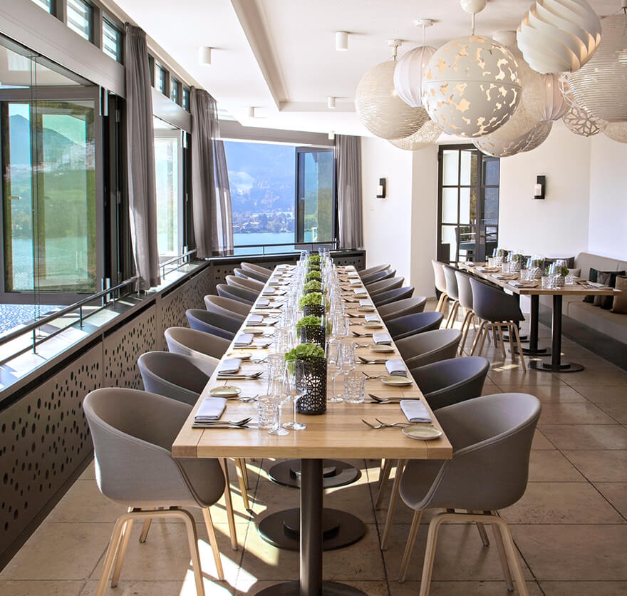 Aufnahme einer langen Tafel mit hellgrauen Polster-Stühlen im Restaurant Alpenbrasserie, die zum bereits eingedeckt ist und sich direkt neben zwei großen und offenen Fenstern mit Blick auf den Tegernsee aufgebaut ist.