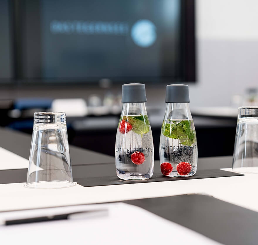 Detailaufnahme von einem Konferenztisch auf dem zwei Gläser und zwei Trinkflaschen, in denen sich Wasser mit frischen Beeren und Minzblättern befindet, stehen.