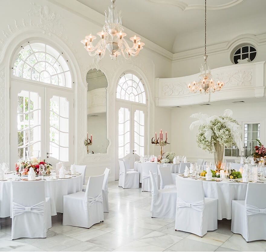 Aufnahme eines Festsaals mit hohen Wänden mit Barockelementen und hellen Bogenfenstern in dem mehrere in weiß und gold eingedeckte Rundtischen und Stühle mit weißen Stuhlhussen stehen.