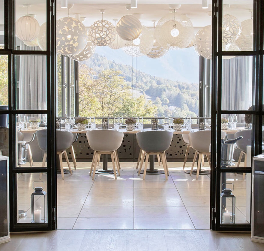 Aufnahme einer langen Tafel in der Eventlocation am Tegernsee im Restaurant Alpenbrasserie, die zum Essen eingedeckt ist und die direkt vor einer langen Fensterfront mit Blick auf die Berge aufgebaut ist.