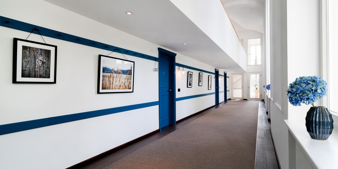 Aufnahme des langen, und hellen Flurs in der Herberge Quirin im Hotel Das Tegernsee mit weißen Wänden und dunkelblauen Farb-Details sowie dunkelblauen Zimmertüren.