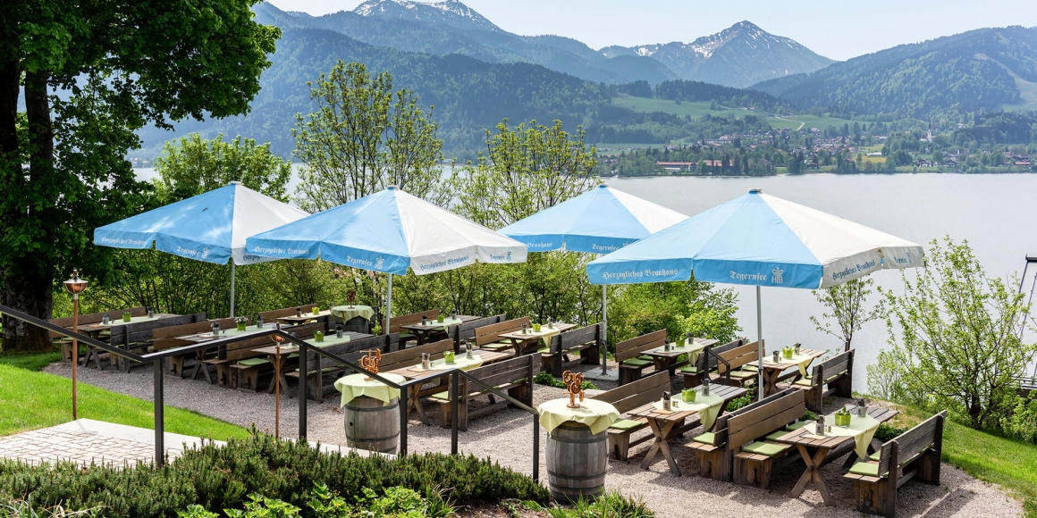 Blick auf den Biergarten auf der Terrasse mit Holzgarnituren und blau-weißen Schirmen, umgeben von Wiese und Bäumen mit Blick auf den Tegernsee und die Berge im Hintergrund.