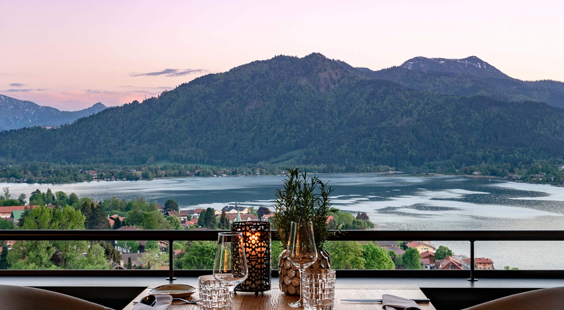 Aufnahme von einem zum Abendessen gedeckten Tisch im Restaurant des Hotels Das Tegernsee mit Blick auf den Tegernsee und die umliegenden Berge während der Abenddämmerung.