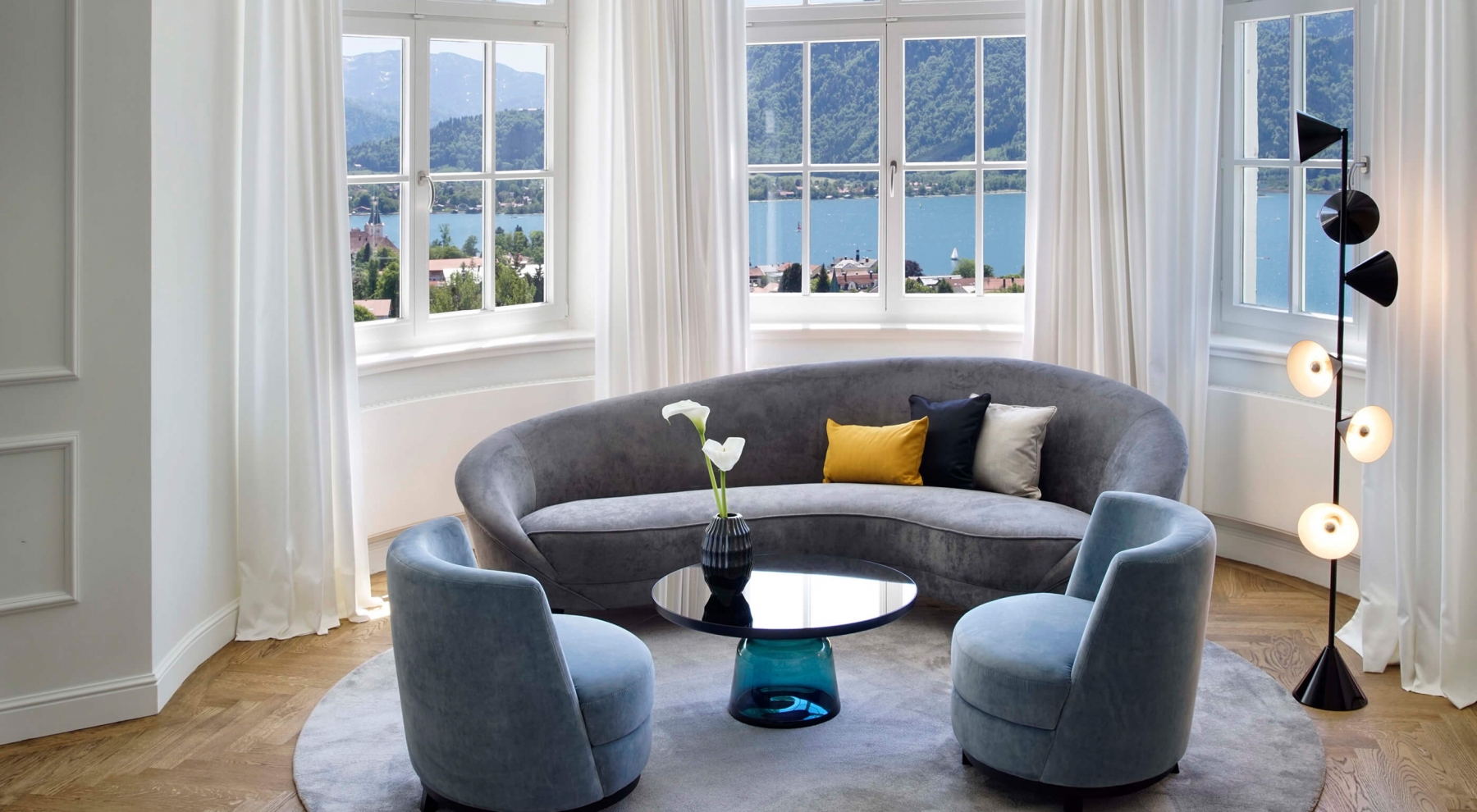 Aufnahme in der Junior Suite im Hotel Das Tegernsee mit einer modernen, hell-grauen Sitzgruppe bestehend aus einem Sofa und zwei Sesseln im Bildzentrum, welche vor einer breiten Fensterfront mit Blick auf den Tegernsee steht.