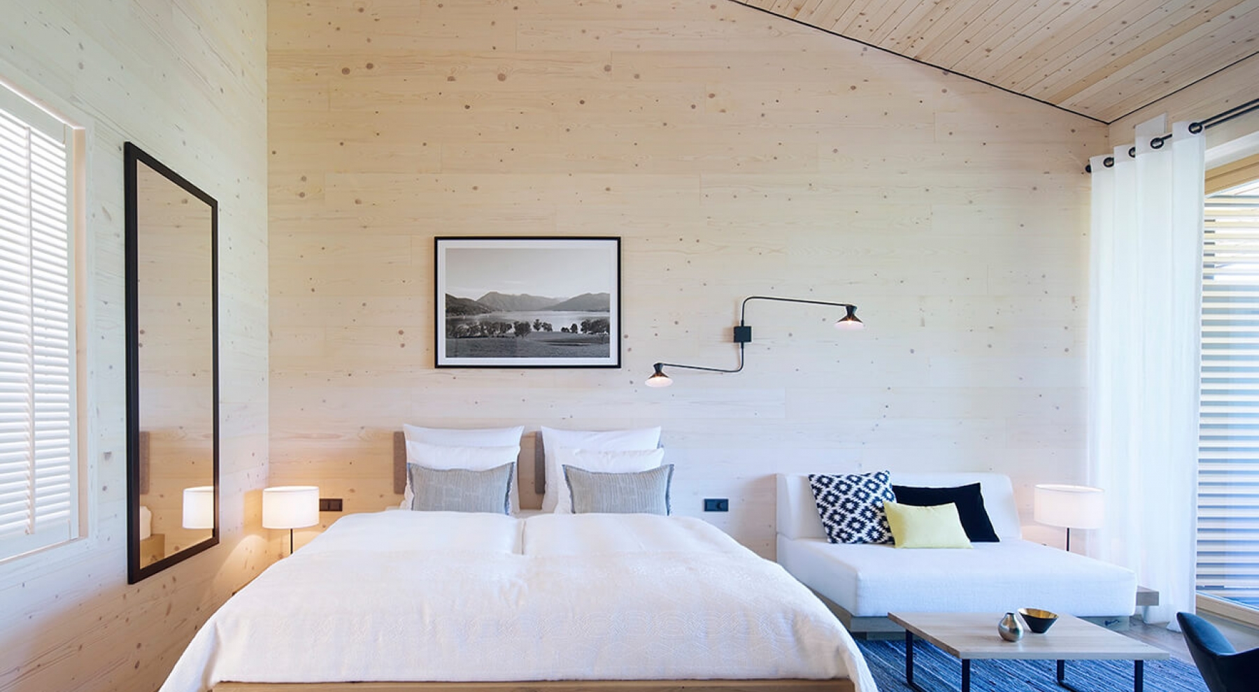 Blick in ein Schlafzimmer im Alpenchalet am Tegernsee mit einem großen Doppelbett, einem weißen Sofa mit Couchtisch aus Holz, einem blau-grauem Teppich und hellen, holzfarben Wänden.