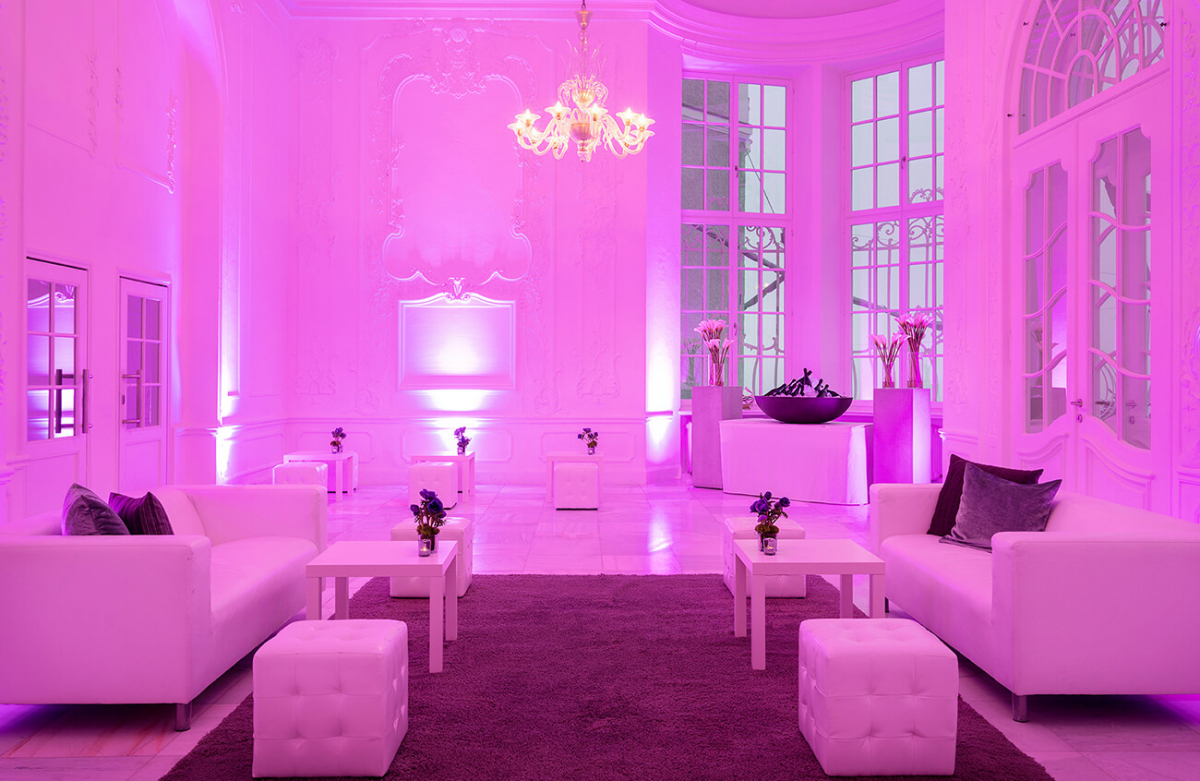 Blick in einen mit rotem Licht ausgeleuchteten Veranstaltungsraum mit hohen weißen Wänden, Lounge-Sofas und Hockern und im Hintergrund eine Getränkebar.