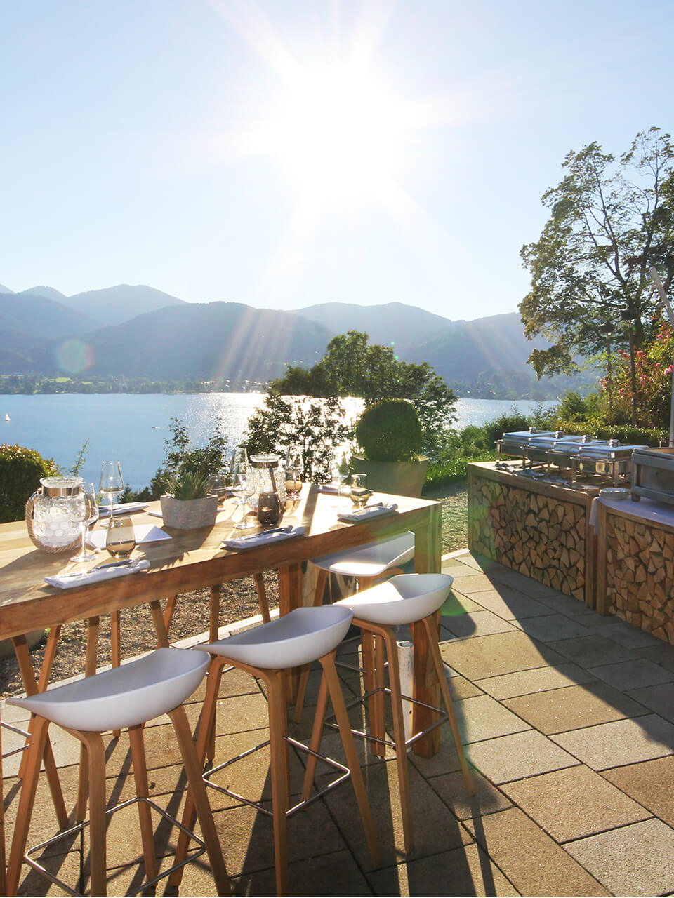 Aufnahme einer Terrasse mit einem hohen Holztisch um den mehrere Barhocker stehen und Panoramablick auf den Tegernsee und die Berge.