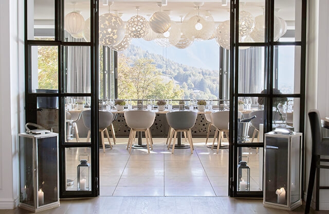 Aufnahme einer langen Tafel im Restaurant Alpenbrasserie, die zum Essen eingedeckt ist und die direkt vor einer langen Fensterfront mit Blick auf die Berge aufgebaut ist.