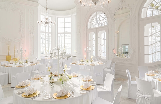Aufnahme eines Festsaals mit hohen Wänden und Bogenfenstern mit mehreren in weiß und gold eingedeckten Rundtischen und Stühlen mit weißen Stuhlhussen.