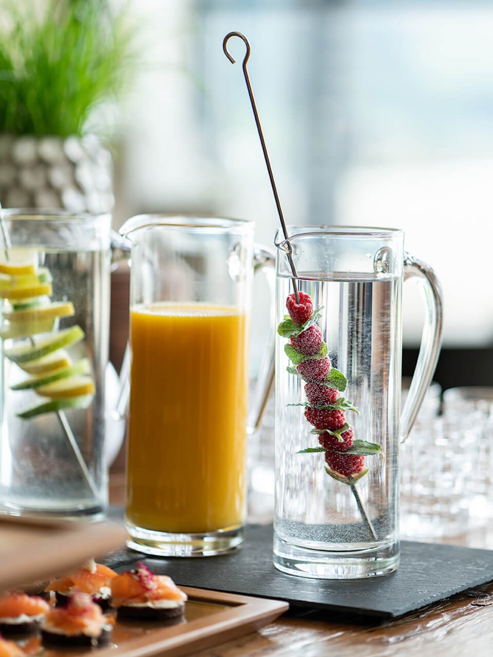 Nahaufnahme eines Buffet-Tisches mit drei Getränkekaraffen mit Orangensaft und Wasser mit frischen Beeren, Minze, Zitrone und Limetten.