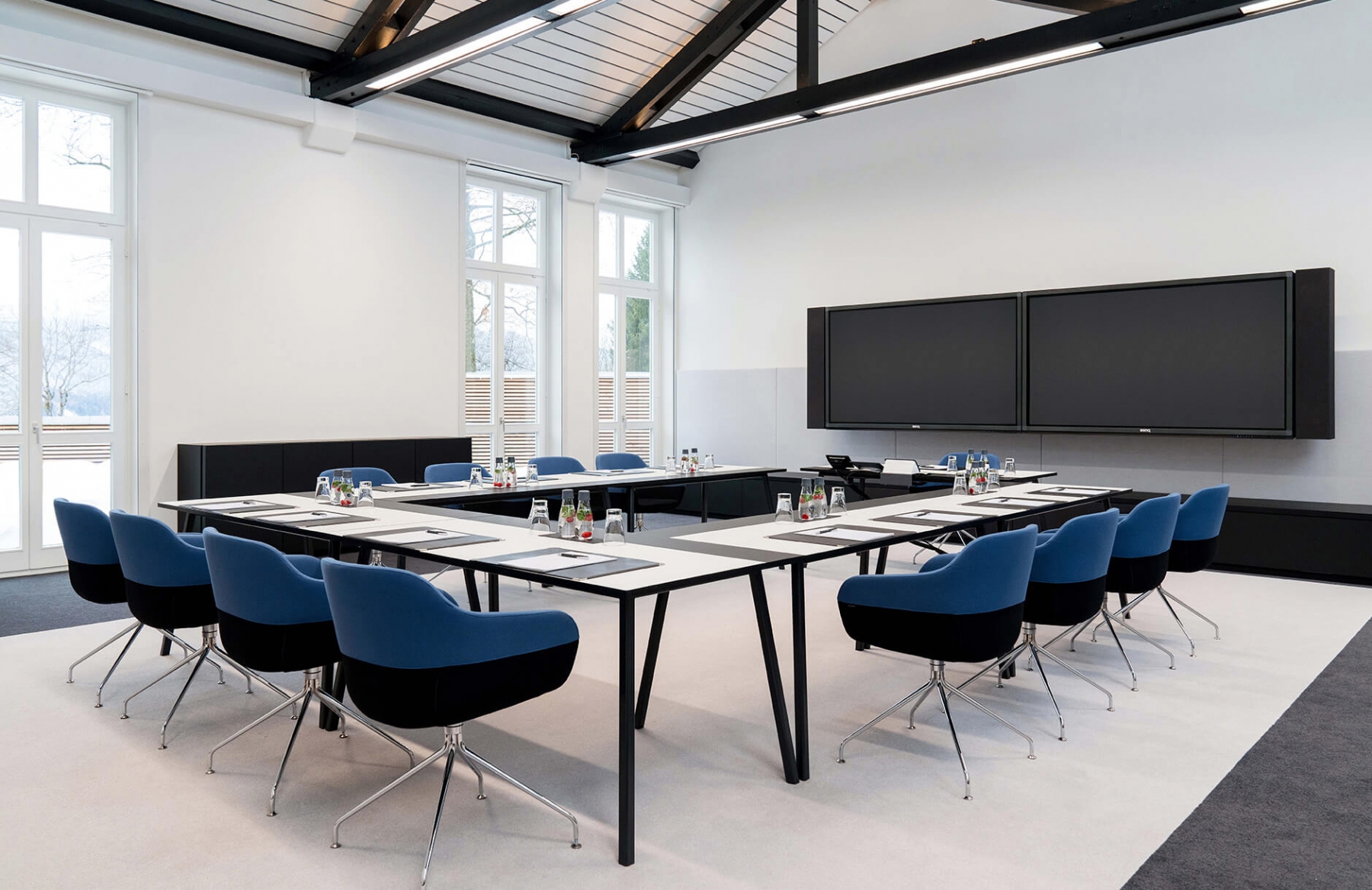 Aufnahme eines Tagungsraumes in dem mehrere Tische und blau-schwarze Stühle in U-Form vor einem schwarzen Bildschirm aufgebaut sind.