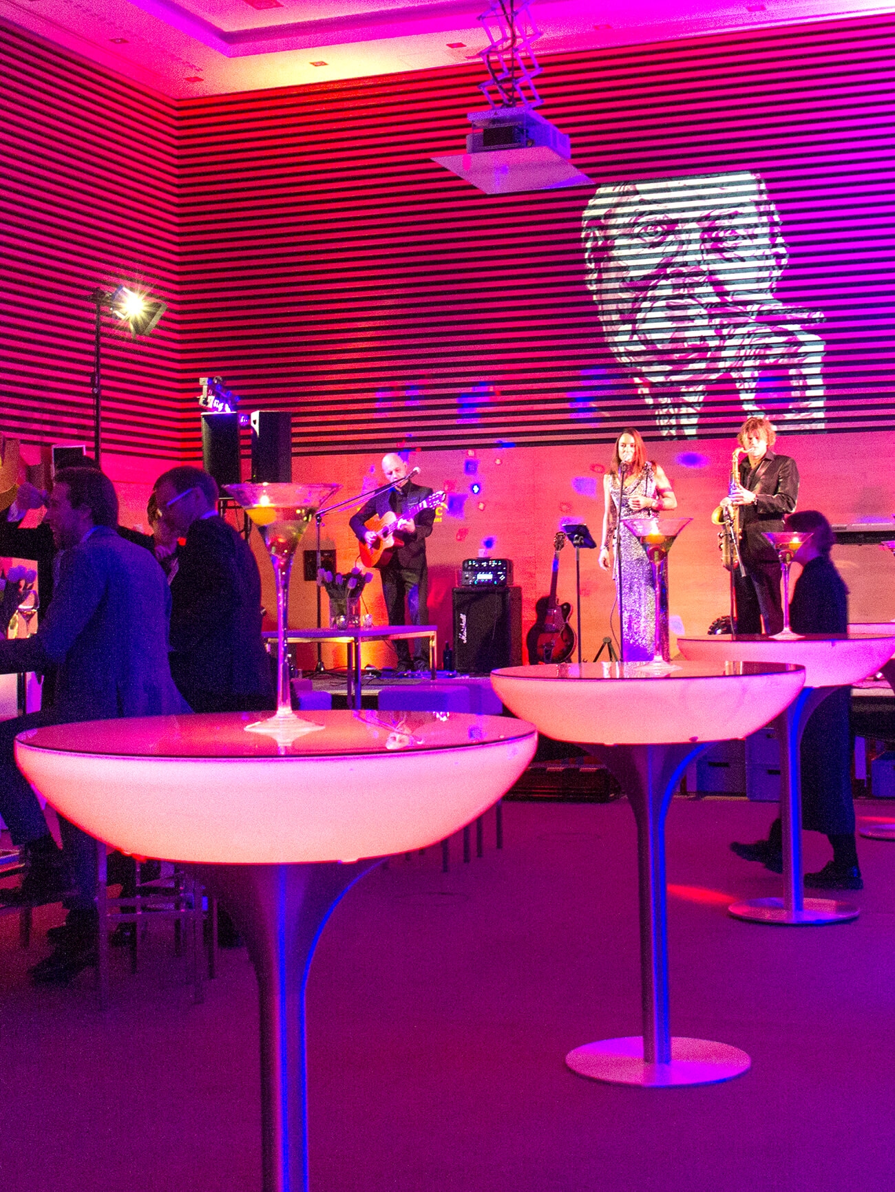 Aufnahme in der Eventlocation am Tegernsee mit einem in roten Lichten beleuchteten Raum, in dem mehrere Stehtische mit Barhockern stehen, um die herum sich mehrere Personen versammeln und im Hintergrund spielt eine Live-Band.